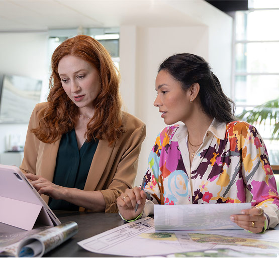 Two women collaborating at a table in a bright, modern office with documents and a computer tablet